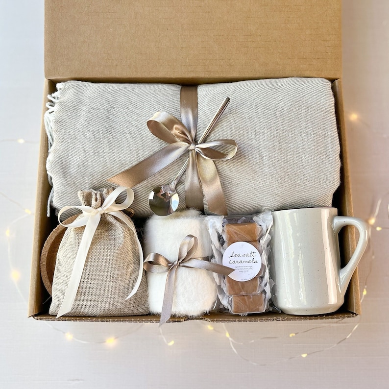 May include: A gift box filled with cosy items for a relaxing evening. It includes a soft grey throw blanket, a mug, a small bag of sea salt caramels, a white fluffy item, and a wooden coaster.