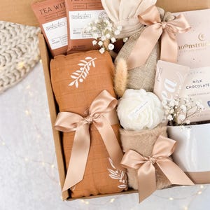 May include: A gift box filled with various items, including tea sachets, a blanket with a white floral design, a candle, and chocolate. The items are tied with satin ribbons in a soft peach color, creating a cohesive and elegant aesthetic.
