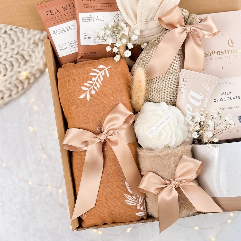May include: A gift box filled with various items, including tea sachets, a blanket with a white floral design, a candle, and chocolate. The items are tied with satin ribbons in a soft peach color, creating a cohesive and elegant aesthetic.