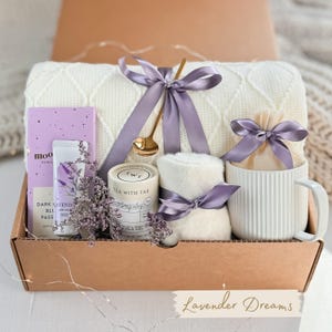 May include: A gift box filled with lavender-themed items, including a white blanket, a candle, a mug, a hand cream, and a tea bag. The box is decorated with a purple ribbon and the words "Lavender Dreams".
