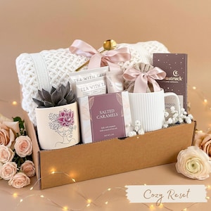 Pu&ograve; includere: Un cesto regalo con una coperta bianca lavorata a maglia, una pianta succulenta in un vaso decorativo, t&egrave;, caramelle al sale, una tazza e una tavoletta di cioccolato. Il cesto &egrave; legato con un nastro rosa e include il testo "Cozy Reset".
