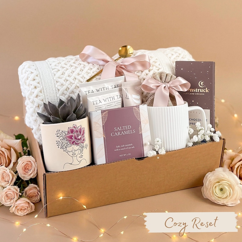 Pu&ograve; includere: Un cesto regalo con una coperta bianca lavorata a maglia, una pianta succulenta in un vaso decorativo, t&egrave;, caramelle al sale, una tazza e una tavoletta di cioccolato. Il cesto &egrave; legato con un nastro rosa e include il testo "Cozy Reset".