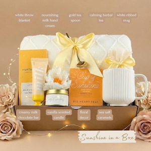 May include: A gift box filled with self-care items. Includes a white throw blanket, mug, tea, hand cream, candle, chocolate bar, and floral decor. The box is tied with a yellow bow and includes the text "Sunshine in a Box".