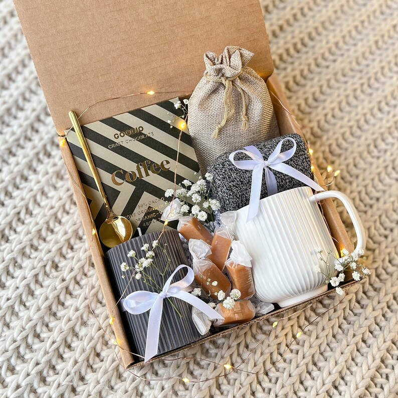 May include: A gift box filled with coffee-themed items. Includes a chocolate bar, a gold spoon, a gray candle, a white mug, a small burlap bag, wrapped candies, and a pair of gray socks tied with white ribbons. The box is decorated with baby's breath.