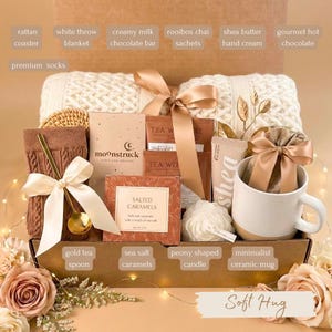 May include: A gift box filled with a white throw blanket, a minimalist ceramic mug, and a gold tea spoon. Other items include a creamy milk chocolate bar, rooibos chai sachets, shea butter hand cream, and sea salt caramels. The box is tied with a ribbon and includes the text "Soft Hug".