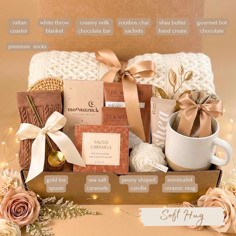 May include: A gift box filled with a white throw blanket, a minimalist ceramic mug, and a gold tea spoon. Other items include a creamy milk chocolate bar, rooibos chai sachets, shea butter hand cream, and sea salt caramels. The box is tied with a ribbon and includes the text "Soft Hug".
