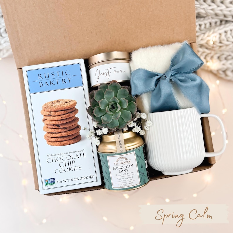 Puede incluir: Una caja de regalo con galletas con chispas de chocolate, una taza blanca, una vela, una suculenta, una lata de t&eacute; y un art&iacute;culo blanco esponjoso atado con una cinta azul. La caja est&aacute; etiquetada como "Rustic Bakery".