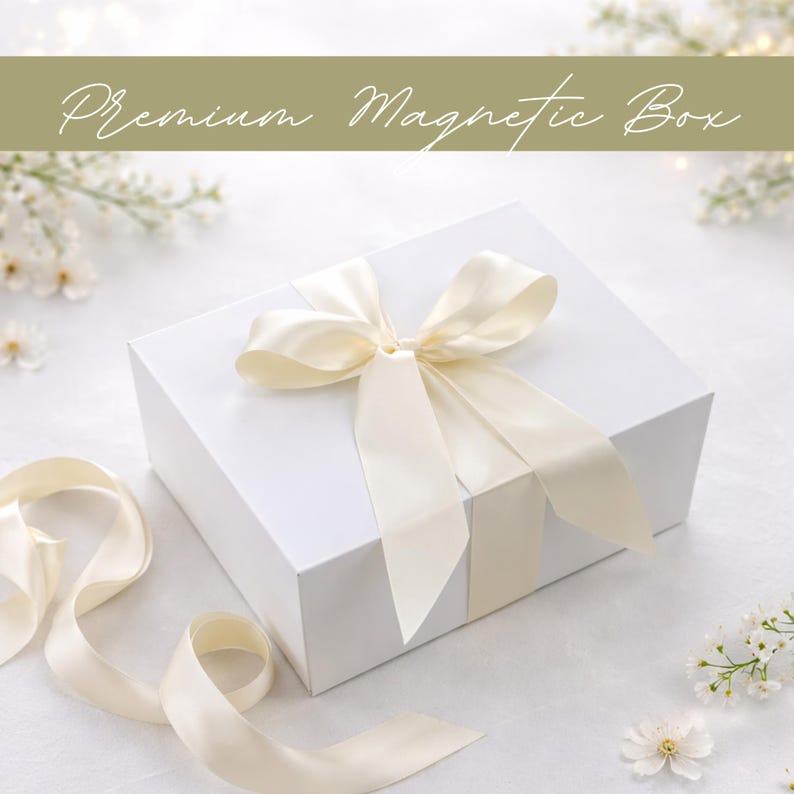 Puede incluir: Una caja de regalo magn&eacute;tica blanca atada con una cinta y lazo de sat&eacute;n color crema. La caja tiene el texto "Premium Magnetic Box" en cursiva. El fondo es blanco con peque&ntilde;as flores blancas y una cinta.