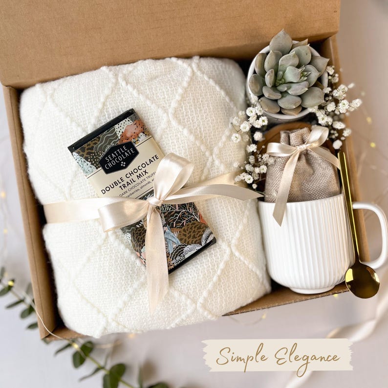 Puede incluir: Una caja de regalo con una manta blanca, una barra de chocolate, una taza blanca, una planta suculenta y una cuchara dorada. La manta est&aacute; atada con una cinta crema. La taza tiene l&iacute;neas verticales. La barra de chocolate est&aacute; etiquetada como "Double Chocolate Trail Mix".