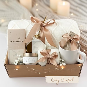 May include: A gift basket filled with cozy items. It includes a white blanket, a mug, a candle, a bar of chocolate, and a small towel, all tied with tan ribbons. The basket has the words "Cozy Comfort" on a banner.