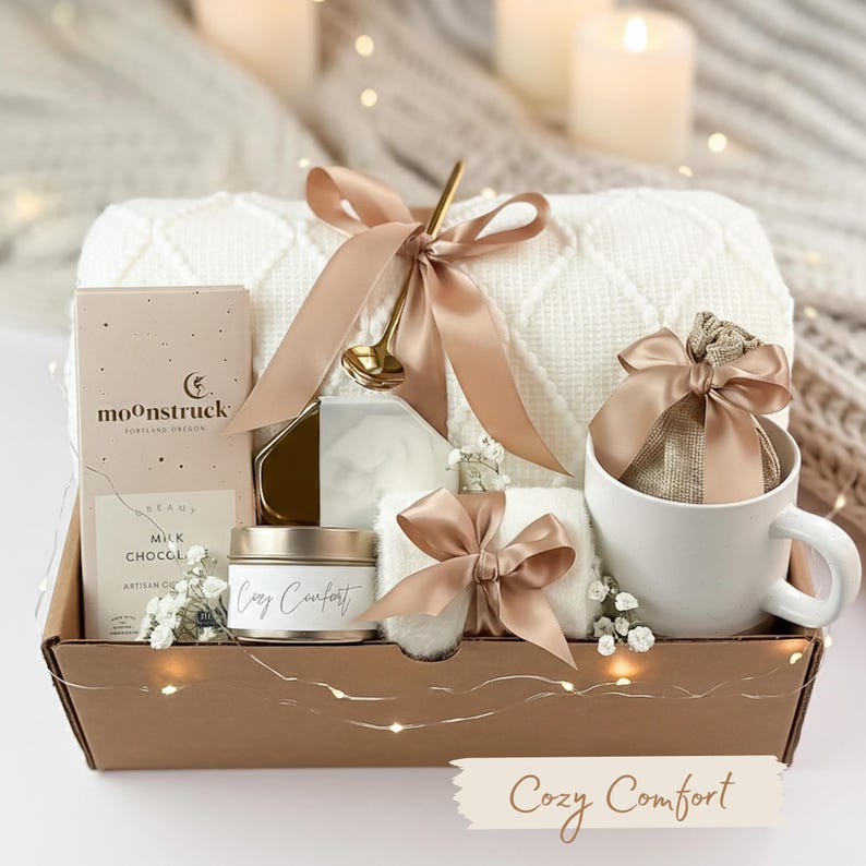 May include: A gift basket filled with cozy items. It includes a white blanket, a mug, a candle, a bar of chocolate, and a small towel, all tied with tan ribbons. The basket has the words "Cozy Comfort" on a banner.