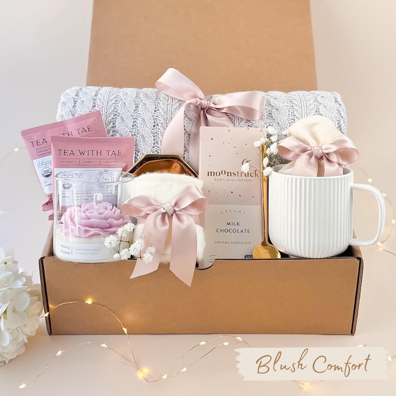 Pu&ograve; includere: Una confezione regalo piena di articoli per la cura di s&eacute;. Include una coperta di maglia grigia, bustine di t&egrave; rosa, una candela, una tazza, una barretta di cioccolato e un oggetto soffice, tutti legati con nastri rosa. La scatola &egrave; etichettata "Blush Comfort".