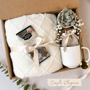 May include: A gift box with a white blanket, a chocolate bar, a white mug, a succulent plant, and a gold spoon. The blanket is tied with a cream ribbon. The mug has vertical lines. The chocolate bar is labeled "Double Chocolate Trail Mix".