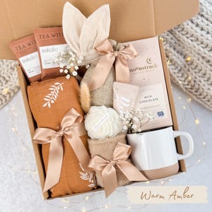 May include: A gift box filled with various items, including tea packets, a muslin blanket with a leaf design, a chocolate bar, a mug, and a decorative flower. The items are tied with satin ribbons. The box is labeled "Warm Amber."