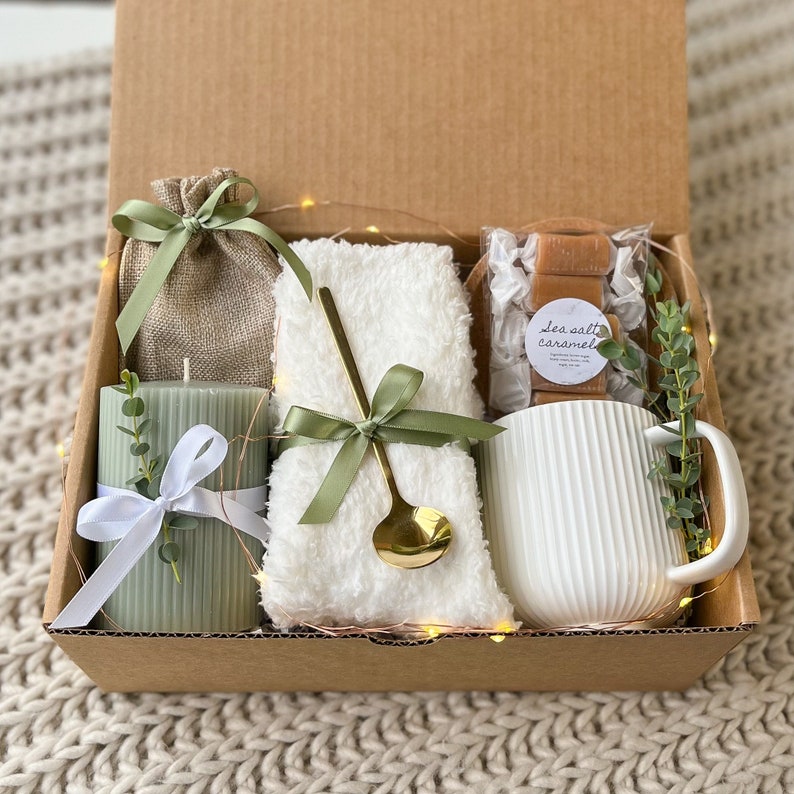 May include: A gift box filled with a white towel, a green candle, a small bag of sea salt caramel sweets, and a white mug. The box is lined with brown paper and has a green and white ribbon.