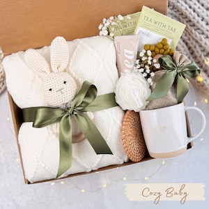 May include: A gift box filled with a white knitted baby blanket, a crochet bunny, a white mug with a heart, tea packets, a candle, and a small woven coaster. The box is tied with a green ribbon. The text "Cozy Baby" is at the bottom.