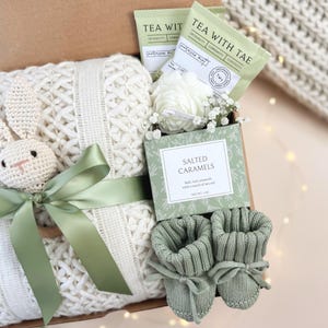 May include: A gift box containing a white knitted blanket, a crocheted bunny, green ribbon, baby booties, tea packets, a box of salted caramels, and white flowers. The box is brown and the overall theme is a baby gift.