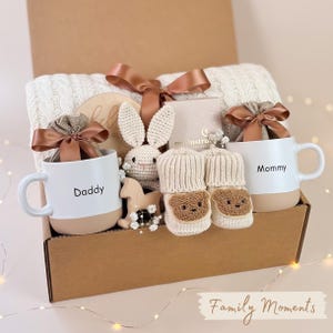 May include: A gift box filled with baby items, including a white blanket, two mugs with "Daddy" and "Mommy" printed on them, a crocheted bunny, baby booties, and a wooden toy. The box is tied with a brown ribbon and has the words "Family Moments" on the bottom.
