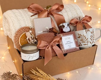 May include: A gift box containing a cream cable-knit blanket tied with a copper ribbon, a candle labelled I AM Joyful, a Moonstruck box, a small honey jar, a brown knit item, a box of Lumi Apple Cinnamon tea, and a mug with a brown leaf pattern.