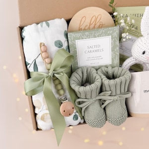 May include: A baby gift box filled with a muslin blanket, pacifier clip, knitted booties, a wooden sign that says "hello", a box of salted caramels, and a white crochet bunny rattle. The items are in shades of green and white.