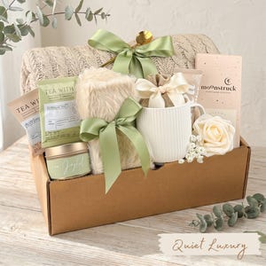 Puede incluir: Una cesta de regalo llena de una manta beige, t&eacute;, una taza, una vela y un art&iacute;culo de piel sint&eacute;tica, todo atado con cintas verde salvia. La cesta incluye una cuchara dorada y una rosa. El texto "Quiet Luxury" est&aacute; en la parte inferior.