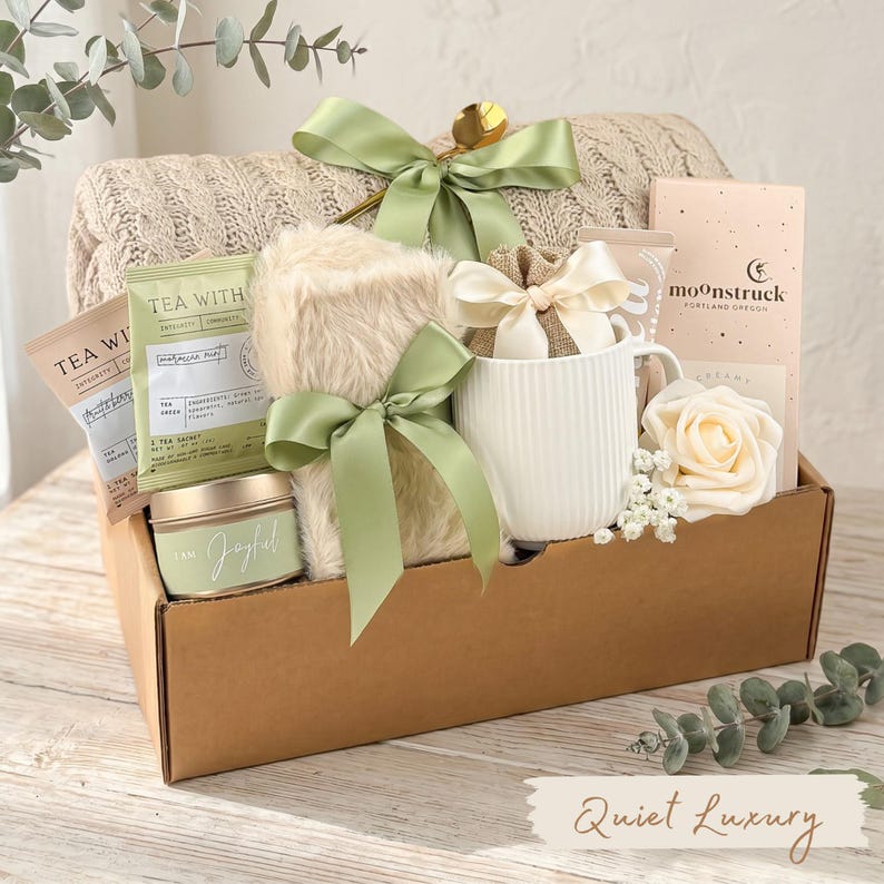 Puede incluir: Una cesta de regalo llena de una manta beige, t&eacute;, una taza, una vela y un art&iacute;culo de piel sint&eacute;tica, todo atado con cintas verde salvia. La cesta incluye una cuchara dorada y una rosa. El texto "Quiet Luxury" est&aacute; en la parte inferior.