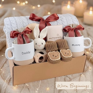 May include: A gift box filled with baby items, including two mugs with "Daddy" and "Mommy" printed on them, a knitted bunny, and bear-shaped baby socks. The box is tied with a burgundy ribbon and includes a blanket and a wooden toy.