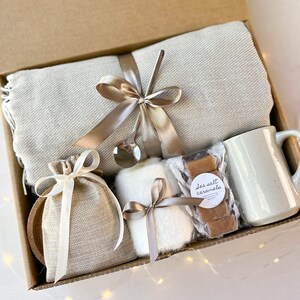 May include: A gift basket filled with cosy items, including a throw blanket, a mug, a small bag, a white fluffy item, and a package of sea salt caramels.