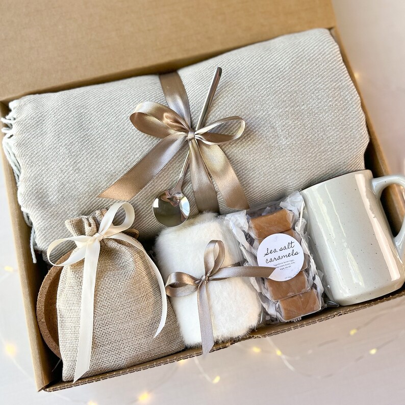 May include: A gift basket filled with cosy items, including a throw blanket, a mug, a small bag, a white fluffy item, and a package of sea salt caramels.