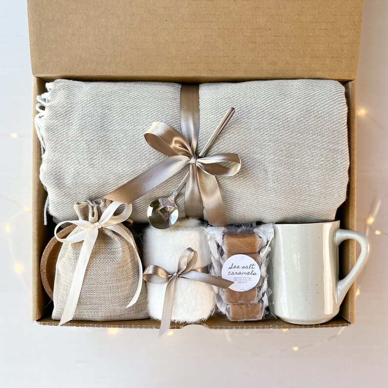 May include: A gift box filled with cosy items, including a grey throw blanket, a white mug, a small bag of sea salt caramels, a white fluffy towel, and a silver spoon.