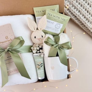 May include: A gift box filled with a white plush blanket, a crocheted bunny, a mug with a heart, tea packets, a tube of hand cream, and a chocolate bar. The box is tied with sage green ribbons.