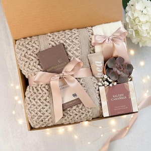 May include: A gift box filled with a beige knit blanket, a bar of chocolate, a tube of lotion, a small succulent plant, and a box of salted caramels. The items are tied with a pink ribbon.