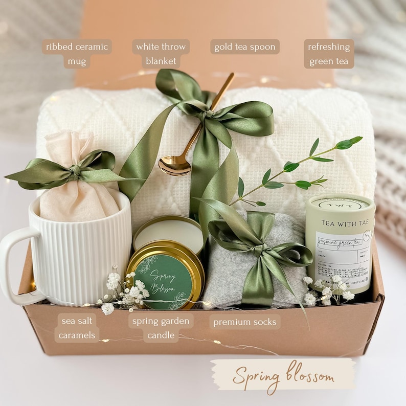 May include: A gift box with a white ribbed ceramic mug, a white throw blanket, a gold tea spoon, and refreshing green tea. The box also contains sea salt caramels, a spring garden candle, and premium socks, all tied with green ribbons. The words "Spring blossom" are written on the box.