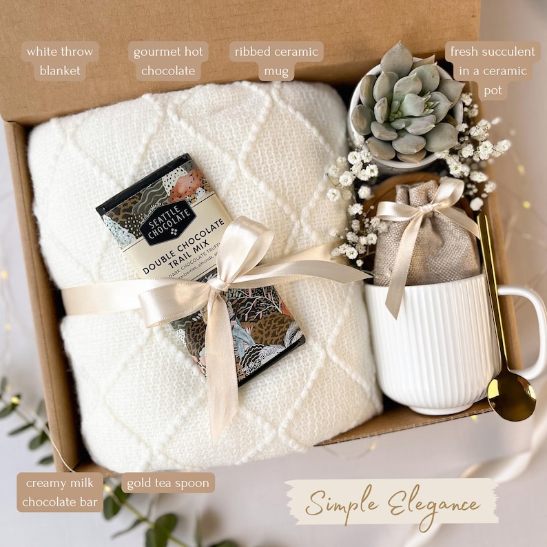 May include: A gift box with a white throw blanket, gourmet hot chocolate, a ribbed ceramic mug, a fresh succulent in a ceramic pot, a creamy milk chocolate bar, and a gold tea spoon. The box is tied with a ribbon and includes the text "Simple Elegance".