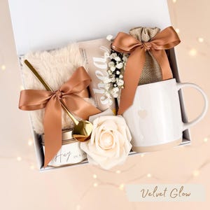 Puede incluir: Una caja de regalo con una taza blanca, una cuchara dorada, una vela, una bolsa peque&ntilde;a y un art&iacute;culo de piel sint&eacute;tica. La taza tiene un dise&ntilde;o de coraz&oacute;n. Los art&iacute;culos est&aacute;n atados con cintas de sat&eacute;n marr&oacute;n y una rosa blanca. La caja tiene el texto "Velvet Glow".