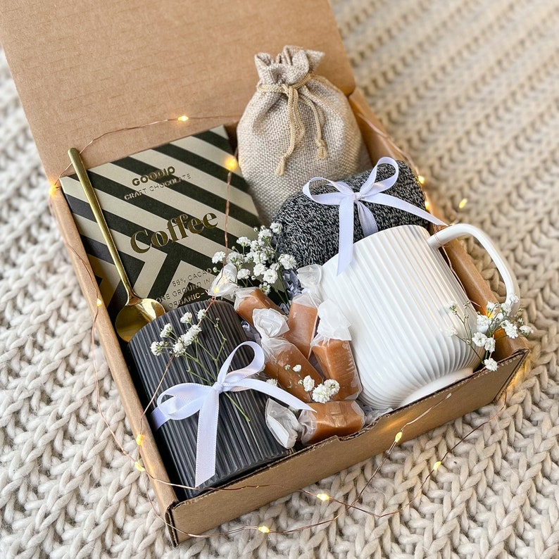 May include: A gift box filled with a mug, chocolate, and other treats. The box contains a white ribbed mug, a dark gray ribbed candle, a gold spoon, a bag of coffee, and wrapped caramels. The box is tied with white ribbons and decorated with small white flowers.