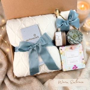 May include: A gift box with a cream-colored cable knit blanket tied with a blue ribbon. The box includes a gold spoon, a small candle, a succulent, and a tea collection box. The text "moonstruck" is visible on a card.