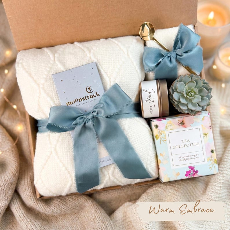 May include: A gift box with a cream-colored cable knit blanket tied with a blue ribbon. The box includes a gold spoon, a small candle, a succulent, and a tea collection box. The text "moonstruck" is visible on a card.