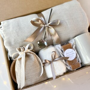 May include: A gift box filled with cosy items, including a soft throw blanket, a small bag of sea salt caramels, a mug, a small bag of what appears to be bath salts, and a small white fluffy item. The box is tied with a brown ribbon.