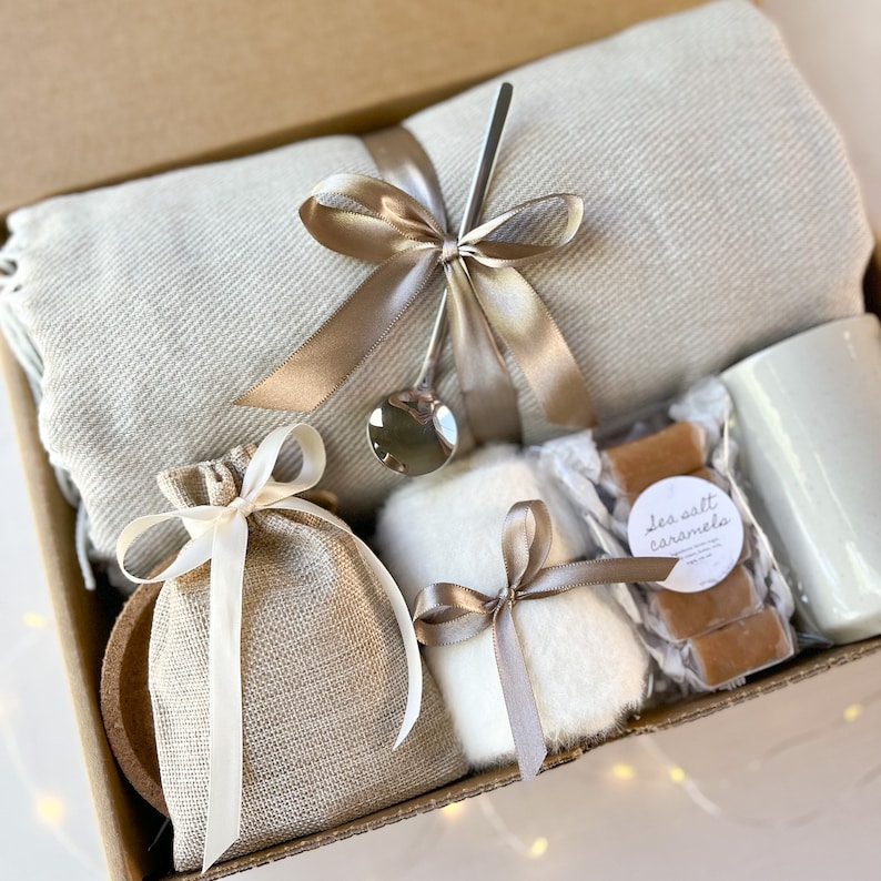 May include: A gift box filled with cosy items, including a soft throw blanket, a small bag of sea salt caramels, a mug, a small bag of what appears to be bath salts, and a small white fluffy item. The box is tied with a brown ribbon.