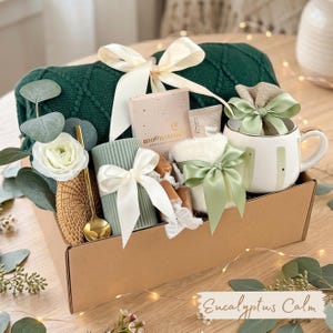 Puede incluir: Una caja de regalo con una manta de punto verde, una taza blanca con detalles dorados, una vela y una caja peque&ntilde;a con el texto "moonstruck". La caja est&aacute; atada con un lazo blanco y el texto "Eucalyptus Calm".