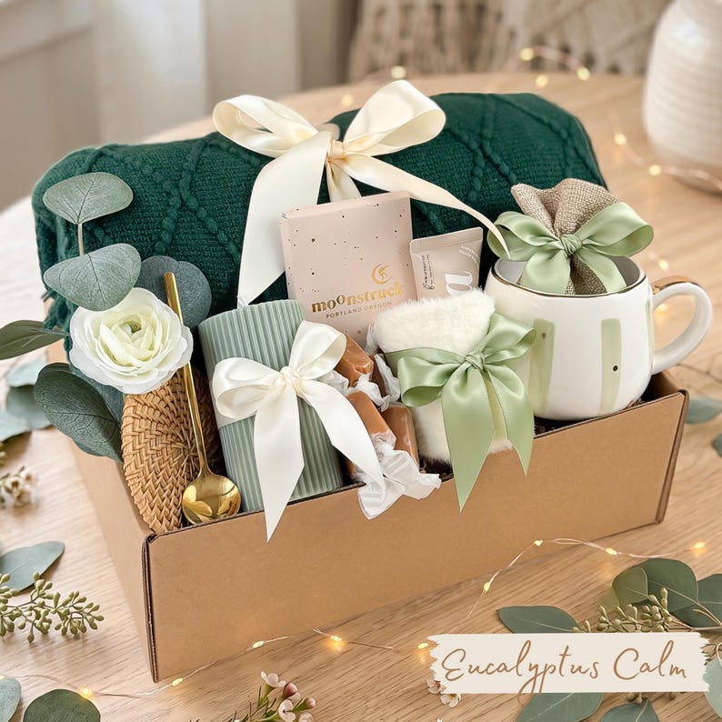 Puede incluir: Una caja de regalo con una manta de punto verde, una taza blanca con detalles dorados, una vela y una caja peque&ntilde;a con el texto "moonstruck". La caja est&aacute; atada con un lazo blanco y el texto "Eucalyptus Calm".