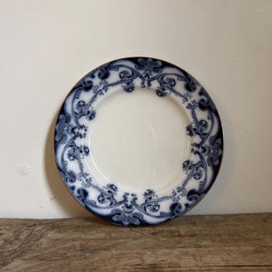 May include: A round ceramic plate with a smooth white center and a wide border featuring a dark blue flowing floral pattern. The edge of the plate is finished with a thin gold-toned rim, creating a distinct contrast against the white surface.