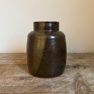 May include: A dark brown ceramic jar with a cylindrical body and a slightly narrower neck. The jar has a textured surface and a reddish-brown base. The jar is sitting on a wooden surface.