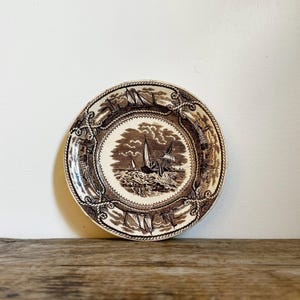 May include: A round ceramic plate featuring a brown and white transferware pattern. The center depicts a sailing ship on turbulent water, while the border displays a nautical rope motif interspersed with additional maritime vessel illustrations.