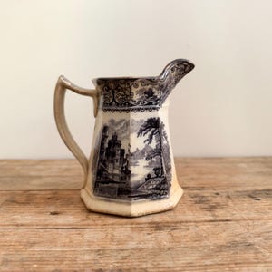 May include: A cream-colored ceramic creamer featuring a dark blue transferware pattern. The pitcher has a hexagonal base, a curved handle, and a spout, decorated with detailed illustrations of a castle, trees, and a landscape scene.