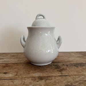 May include: A light gray ceramic sugar bowl with a matching lid. The bowl has two small handles on the sides and a loop handle on the lid. The sugar bowl is sitting on a wooden surface.