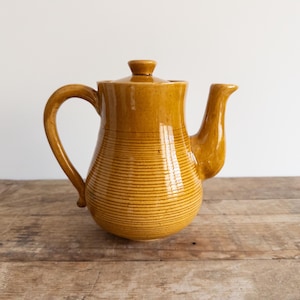 May include: A yellow ceramic teapot with a striped design. The teapot has a rounded body and a spout on the side.