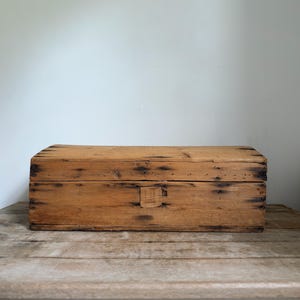 May include: A rectangular wooden box with a natural light brown finish, featuring visible wood grain, dark knots, and small nail heads along the edges. The box has a hinged lid and a small, centered rectangular wooden latch on the front panel.