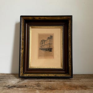 May include: A framed etching depicting a street scene with multi-story buildings, housed in a dark brown wooden frame with gold-toned accents and a cream-colored mat. The artwork features detailed architectural lines and shading.